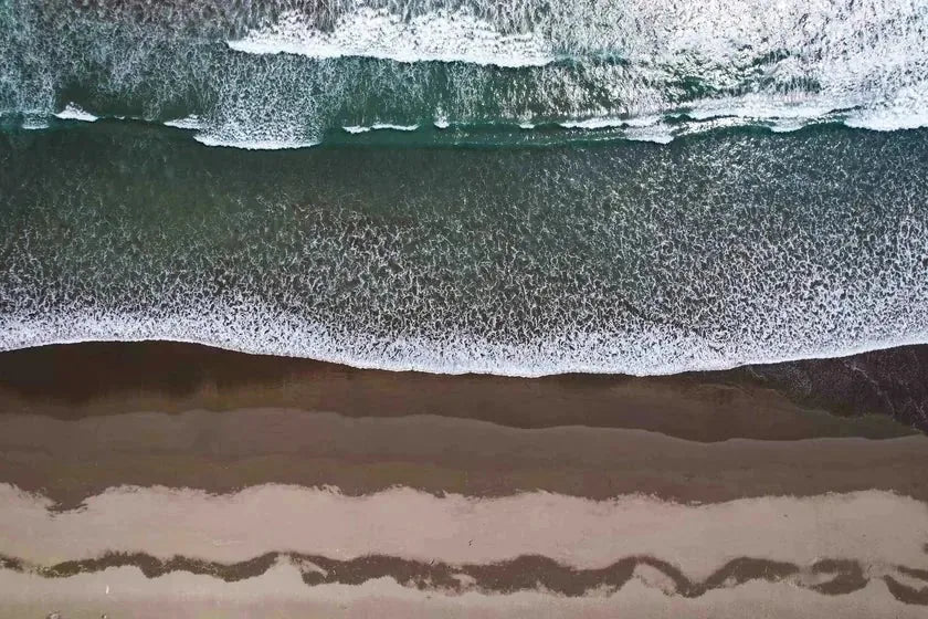 An aerial view of pale sand and foam meeting open water — the lived-in coast as pattern, not place.