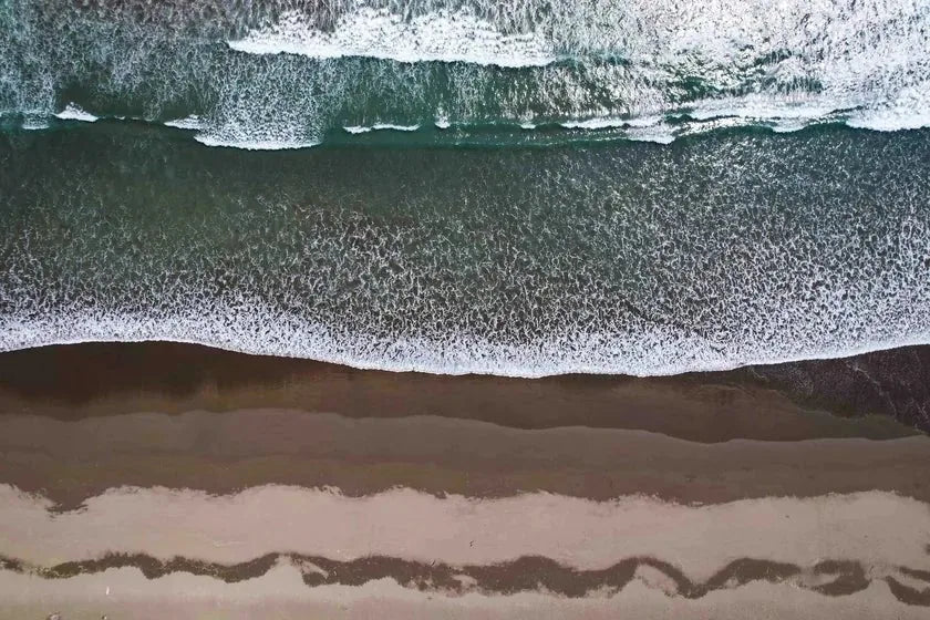 An aerial view of pale sand and foam meeting open water — the lived-in coast as pattern, not place.